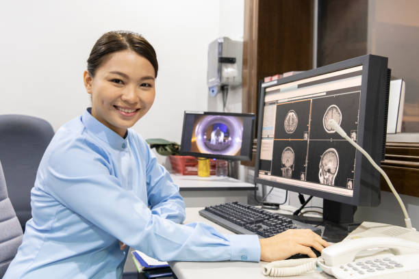 Female nurse, in uniform sitting down at her desk looking confidently at camera with MRI scan medical imaging on her screen